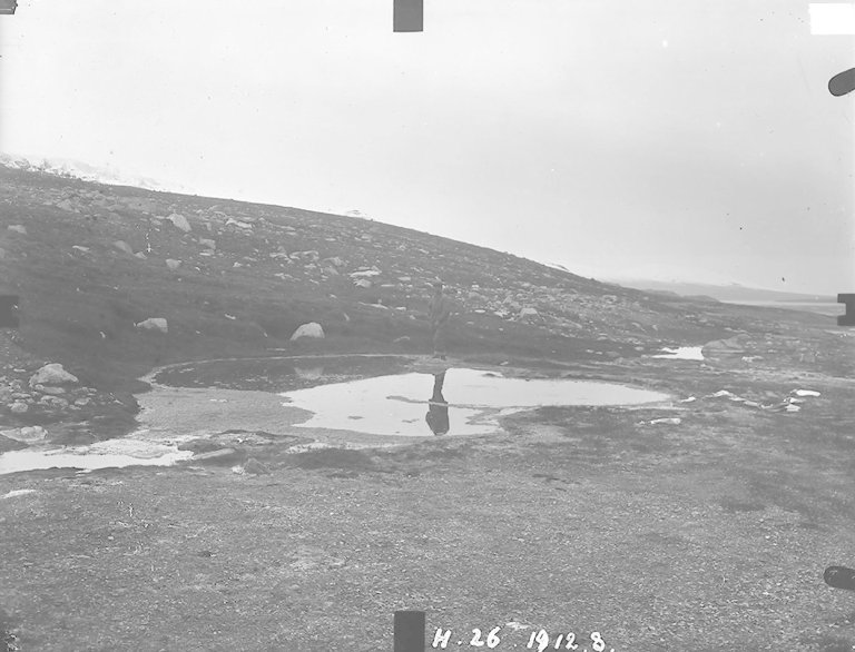 Svart-hvitt bilde av person som står ved lite vann/kilde.