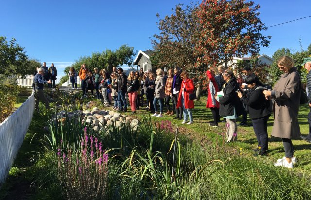 20 onlookers watching Bent Baskerud presenting bluegreen solutions. He is standing against a fence, while they are stand on a path in front of a house.