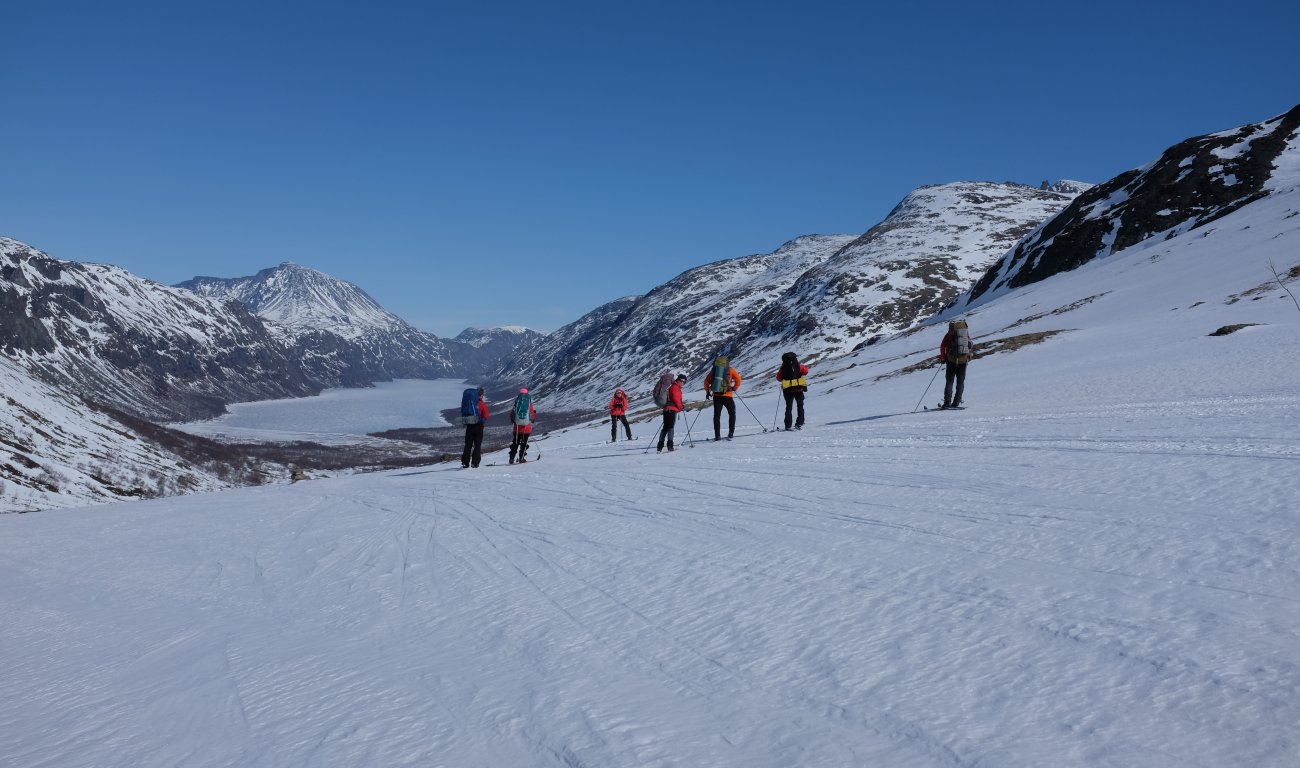 En gruppe skiløpere på vei ned en snødekt bakke. Islagt vann i bakgrunnen. 