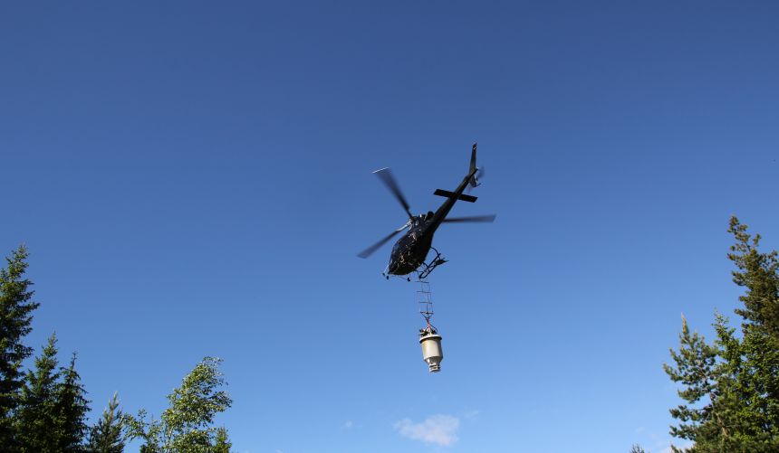 Bilde av helikopter med tønnelignende gjenstand hengende under. 