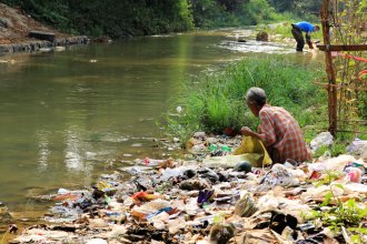 Bilde av person som sitter langs elv med store mengder plastavfall.